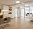 Modern office space with a large wooden table, several chairs, a laptop, a small kitchen area with a coffee machine, and a seating area with a round table and armchairs.