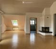 Bright, empty room with wooden flooring, white walls, and a skylight window. Features a built-in fireplace and a visible kitchen area in the background.