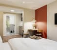 Modern bedroom with a large bed, a small desk with chairs, a vase with flowers, a television mounted on the wall, and an adjacent bathroom with a glass shower enclosure.