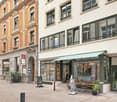 Two adjacent buildings in an urban setting, one with a modern facade and the other with a classic brick design. The modern building has a storefront with a striped awning, outdoor seating, and signage for "Stay Sunday". The classic building features arche