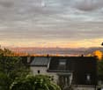 View of a cityscape with a house in the foreground, surrounded by trees, with mountains and a sunset in the background.