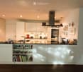 Modern kitchen with white cabinets, black countertops, and various appliances including a sink, fridge, and oven. Shelves display glasses, bowls, and books.