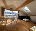 Bright, modern living room with wooden floors, large windows offering a mountain view, a sofa, coffee table, dining table with chairs, and a hanging lamp.