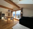 Bright, open living and dining area with wooden flooring, large windows offering a mountain view, a black sofa, a wooden dining table with white chairs, and a modern kitchen area with cabinets.