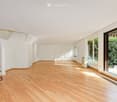 Bright, unfurnished room with wooden flooring, white walls, large windows, and a staircase leading to another level.