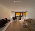 Living room with a sofa, coffee table, TV, dining table, and chairs. Large windows with orange blinds lead to a balcony.
