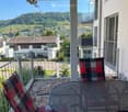 Balcony with a scenic view of a town and hills, featuring a round metal table and two plaid-patterned chairs.