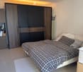 Modern bedroom with a large bed, dark grey wardrobe, nightstand, and a mirror. The room features a white tiled floor and a round ceiling light.