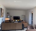 Modern living room with a brown leather sofa, a wooden side table, a lamp, a television, a mirror, and a cabinet. The room features tiled flooring and a slanted ceiling.