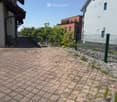 A paved outdoor area with a brick-patterned ground, surrounded by a green metal fence. There are some small plants and shrubs near the fence, and a few buildings are visible in the background under a clear blue sky.