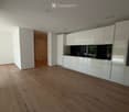 Empty room with light wooden flooring, white walls, and a white kitchen area with cabinets and a countertop visible in the background.