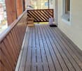 Wooden balcony with slatted flooring, wooden railing, and a wooden pergola overhead. There is a stack of firewood and a small white container on the balcony.