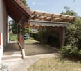 A covered outdoor patio area with wooden beams and a striped awning, surrounded by greenery and a brick pathway leading to a grassy area.