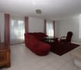 Living room with red sofa, wooden coffee table, red armchair, and wooden cabinet. The room features tiled flooring, large windows with curtains, and a chandelier.
