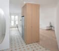 Modern, open-plan living space with a white sofa, a small round table, a large mirror, a refrigerator, and a wooden cabinet. The floor features patterned tiles in the hallway and wooden flooring in the living area. A balcony is visible through large windo