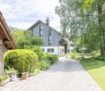 A gravel pathway leads to a cluster of buildings surrounded by lush greenery and well-maintained hedges. The scene includes a wooden shed, a modern house with grey siding, and various potted plants along the pathway.