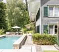 Two-story house with grey shingled roof, white trim, green shutters, and a covered porch. Adjacent to a swimming pool with a stone deck, outdoor seating area, and lush greenery.