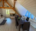 Loft-style room with wooden beams, fireplace, sofa, dining table with chairs, and a fan. Large windows provide natural light.