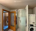 Bathroom with a glass-enclosed shower, a washing machine, and a wooden door leading to another room.