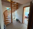 Spacious hallway with wooden flooring, a wooden spiral staircase leading upwards, and a small metal table with items on it. A doorway leads to another room with visible furniture and items.