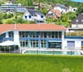 A modern two-story house with a red-tiled roof, large windows, and a swimming pool in the front yard. The house features a garage and is surrounded by lush greenery and neighboring houses.