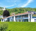 Modern two-story house with large windows, red-tiled roof, and a blue awning, situated on a hillside with a well-maintained lawn and surrounding greenery.