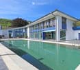 Modern two-story house with large windows, a swimming pool, and a garage. The house features a sleek design with white and blue accents, surrounded by greenery and a clear blue sky.