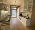 Modern kitchen with white cabinets, black countertops, and a wooden floor. Features a sink, faucet, oven, and a bottle on the counter. A large window with a view of a balcony is visible.