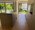 Modern kitchen with light wooden flooring, grey cabinets, and a central island with a black countertop. Large windows and sliding doors lead to a balcony with greenery visible outside.