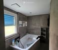 Modern bathroom with a large bathtub, toilet, shelf, and window offering a scenic view. The room features grey tiles, a sleek design, and decorative picture frames on the wall.