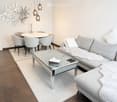 Modern living room with a grey sectional sofa, glass coffee table, dining table with four chairs, and decorative wall art.