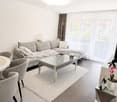 Modern living room with a grey sofa, glass coffee table, two armchairs, a white dining table, and a television. The room features large windows with sheer curtains and a view of the outside.