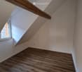 Empty attic room with wooden flooring, white walls, exposed wooden beams, and a small window letting in natural light.