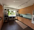 Modern kitchen with light wood cabinetry, dark tiled floor, island with stools, and large windows overlooking a green area.