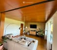Modern living room with wooden flooring, a large sectional sofa, coffee table, TV mounted on a brick wall, floor lamp, and potted plants.