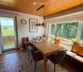 Bright, modern dining area with a wooden table, four chairs, a built-in sofa, pendant lights, a clock, and a picture frame on the wall. Large windows offer a scenic view of greenery.