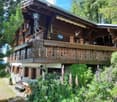 A two-story wooden house with a balcony, decorative wooden railings, and lush garden in front.