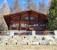 A two-story wooden house with a stone foundation, multiple windows, and a balcony overlooking a natural landscape.