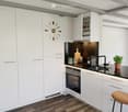 Modern kitchen with white cabinets, dark countertops, and wooden flooring. Features include an oven, sink, coffee maker, and a decorative clock on the wall.