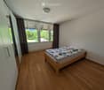 Minimalist bedroom with wooden flooring, a single bed with a colorful quilt, and a large window with curtains offering a view of greenery.