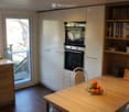modern kitchen with wooden countertops, wooden cabinets, white appliances, sink with faucet, window with glass door, round table with bowl of fruit, and bookcase with books