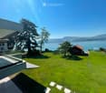 Lush green lawn with a modern glass balcony, a small red shed, and a scenic view of a lake and mountains in the background.