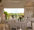 Elegant restaurant interior with large windows overlooking a scenic countryside view, featuring white tablecloths, dining tables, chairs, wine bottles, flower vases, and table lamps.