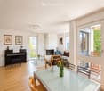 Bright, open living and dining area with wooden flooring, large windows, and a balcony. Features a piano, sofa, armchair, fireplace, glass coffee and dining tables, and chairs.