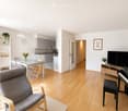 Bright, open-plan living and dining area with light wooden flooring, a grey sofa, a dining table with chairs, a piano, a TV, and a small kitchen area with cabinets and appliances.