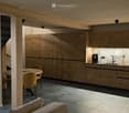 Open-plan living and kitchen area with modern wooden cabinets, a round dining table with chairs, a sofa, and various kitchen appliances including a coffee maker and teapot.