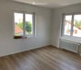 Empty room with white walls, wooden ceiling, and two windows offering views of the outside. Radiator visible on one wall.