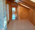 Empty hallway with wooden flooring and walls, featuring a mirror on one side and a view into another room through an open doorway.