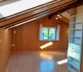 Empty attic room with wooden walls and floor, white built-in shelving unit, skylight window, and exposed wooden beams on the ceiling.