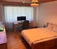 Cozy bedroom with a wooden bed, desk with a computer, chair, and a small fish tank on a side table. The room features wooden flooring and a hanging woven light fixture.
